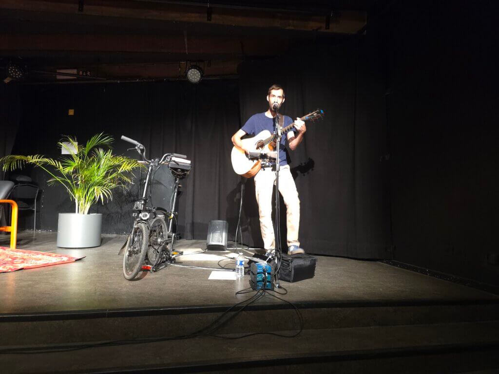 Le chanteur Antoine Armedan, sur scène avec sa guitare et son vélo pliant qui lui sert de support à la console portable.